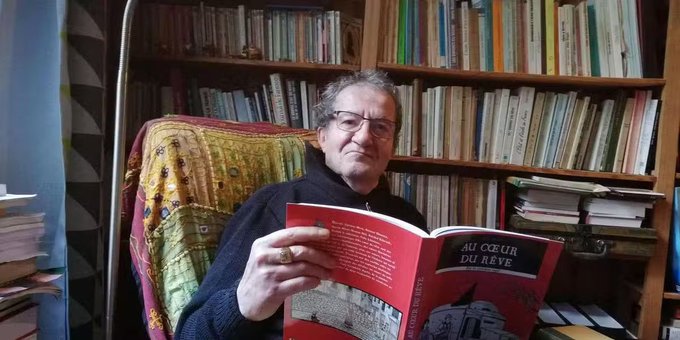 The image shows an older man sitting comfortably in what appears to be a home library or study. He is holding and reading a book with a red cover titled “Au cœur du rêve.” He is wearing glasses and a dark sweater, and has short, graying hair. His expression is neutral to mildly engaged, as if he paused reading to look at the camera.
Behind him are wooden bookshelves filled with many books arranged vertically, creating a dense, academic or literary atmosphere. He is seated in a cushioned chair draped with a colorful, patterned textile. A tall floor lamp stands to the left, and there are additional stacks of books nearby, reinforcing the sense of a personal reading or study space.